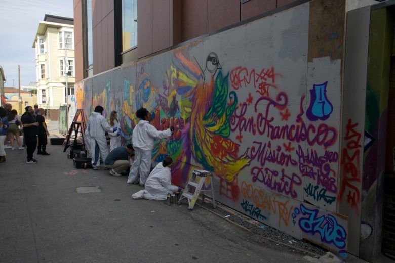 A group of people in white coveralls paint a colorful mural on a wall in an urban alley, with spray paint cans and ladders nearby. Several others observe the scene.
