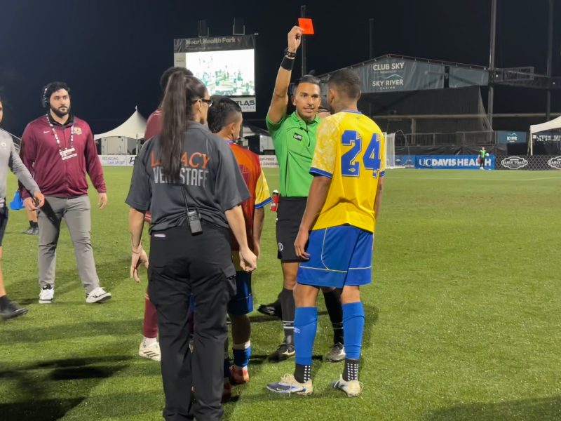 A soccer referee holds up a red card to a player in a yellow jersey on a field at night while other players and staff stand nearby.