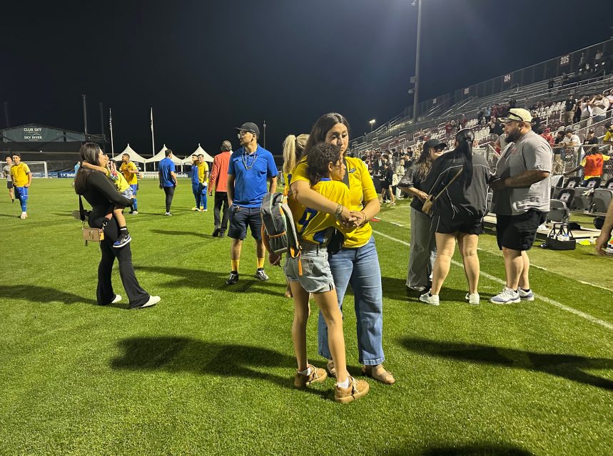 People gather on a soccer field at night; some are talking, hugging, or standing, while others walk in the background near stadium seating.