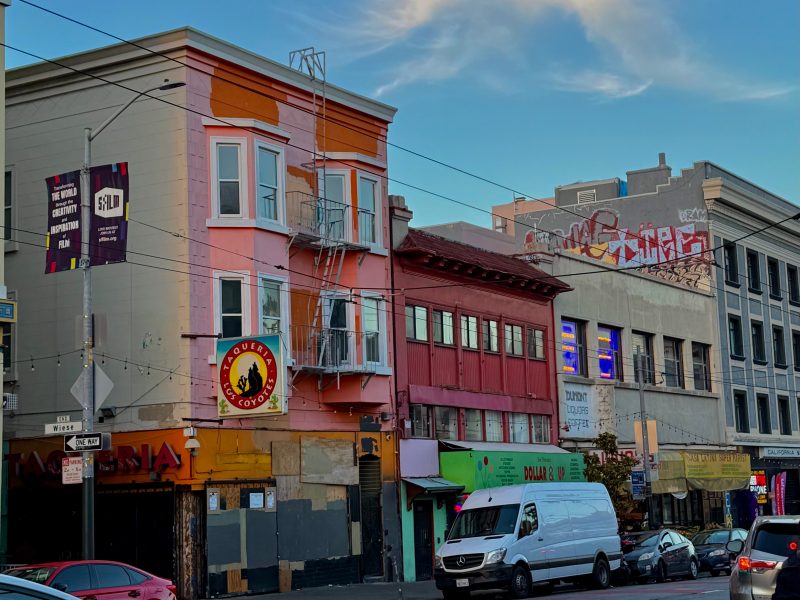 Street view of colorful buildings, including a pink and orange corner building with a "Queen's Co" sign, a white van parked in front, and graffiti on a rooftop in the background.