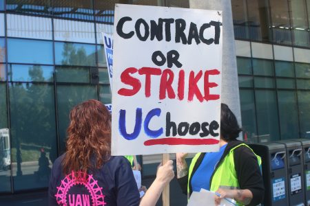 Two people hold a protest sign that reads “Contract or Strike UCchoose” outside a building; one wears a shirt with a UAW logo, the other a yellow vest.