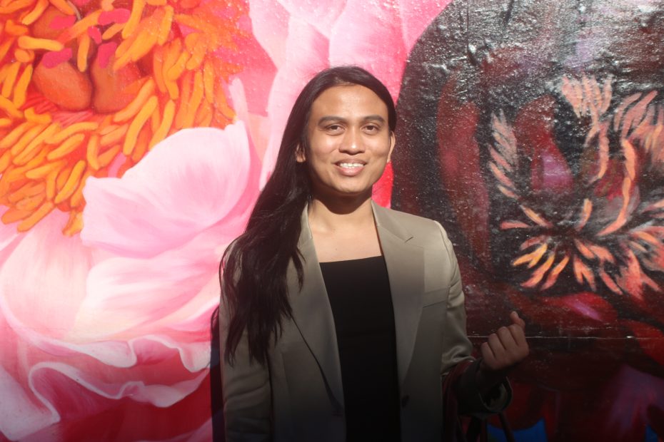 A person with long dark hair wearing a beige blazer stands in front of a colorful mural featuring large pink and orange flowers.