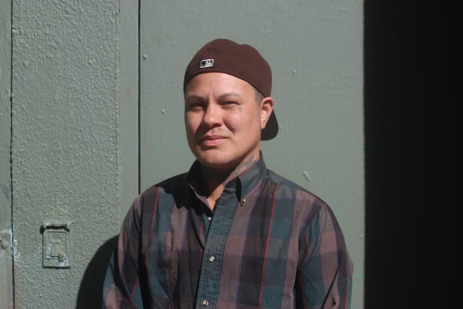 Person wearing a brown cap and plaid shirt stands in sunlight against a textured green wall, partially shaded.