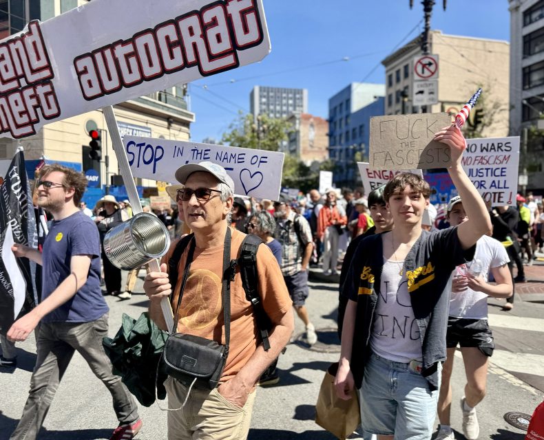A group of people march in a protest, holding signs with messages against autocracy, fascism, and war, on a city street in daylight.
