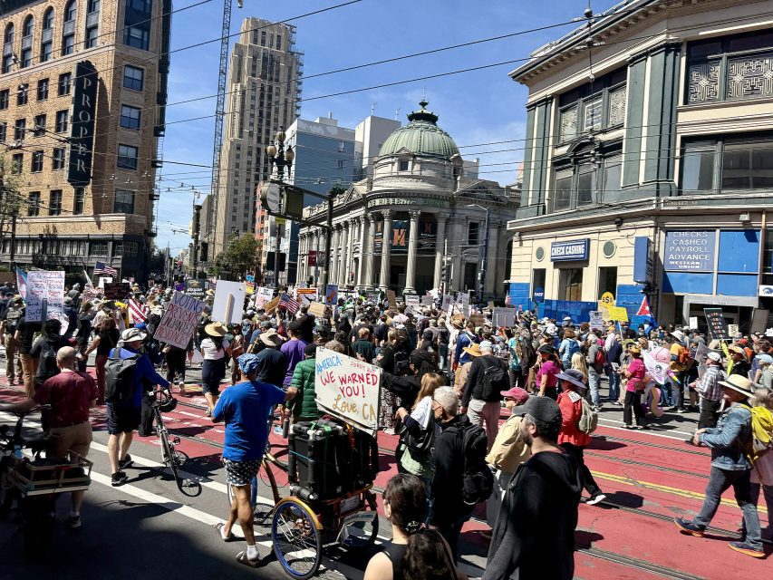 A large crowd of people participates in a protest on a city street, holding signs and banners, with tall buildings visible in the background.