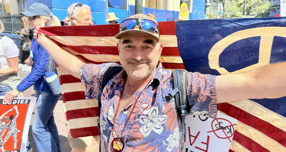 A man wearing a floral shirt and hat stands with arms outstretched, holding an American flag, in a crowd at a protest. Signs with red-circled slashes are visible.
