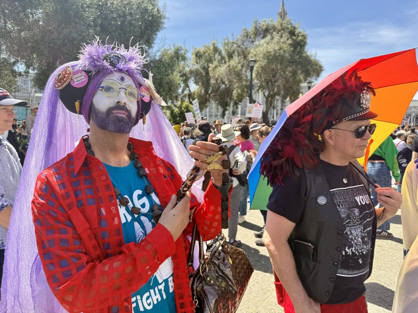 Two people in colorful, festive outfits stand outdoors at a crowded event; one holds a religious cross, and the other holds a rainbow umbrella.