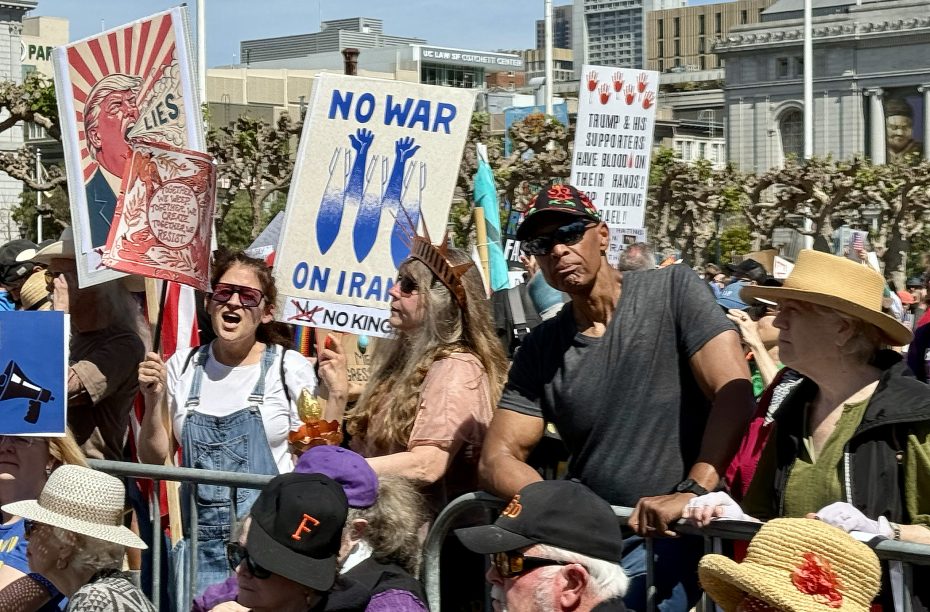 A crowd of people at a protest holds signs with messages against war and political figures; some wear hats and sunglasses on a sunny day in an urban setting.