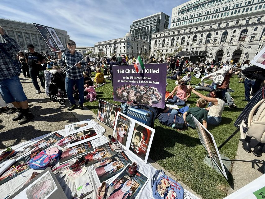 A public protest displays signs and portraits on the grass, including one with the message "168 children killed in the US-Israel strike on an elementary school in Iran.
