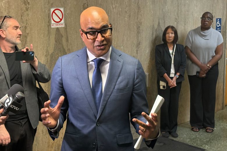 A man in a suit speaks to the media inside a building, holding papers, while several people stand in the background.