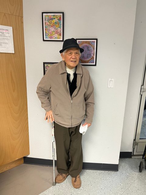 An elderly man wearing a hat and jacket stands indoors with a cane, holding a paper, in front of a wall with colorful framed artwork.