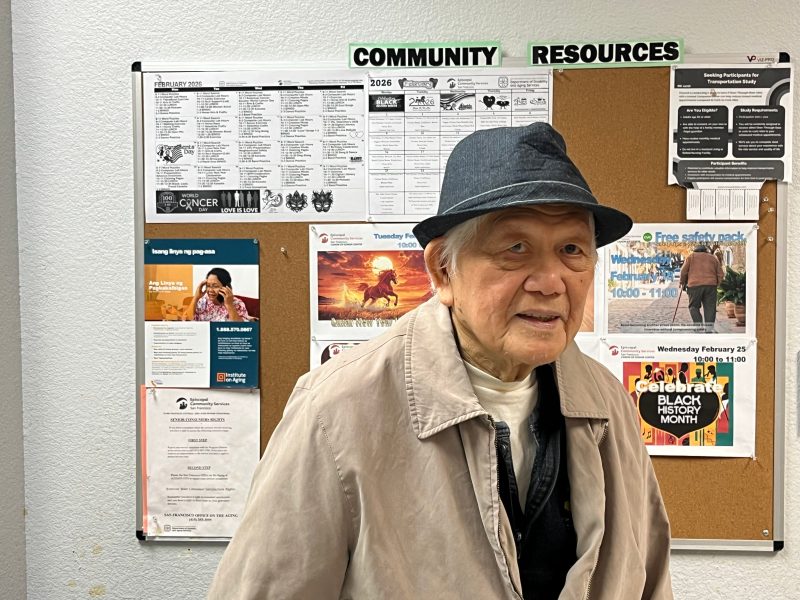 An elderly man in a hat and coat stands in front of a bulletin board labeled "Community Resources" with various flyers and notices posted.
