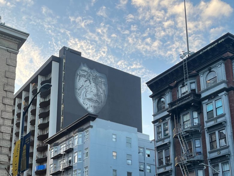 Large mural of a stylized face with intricate patterns on the side of a tall building, surrounded by other buildings under a partly cloudy sky.