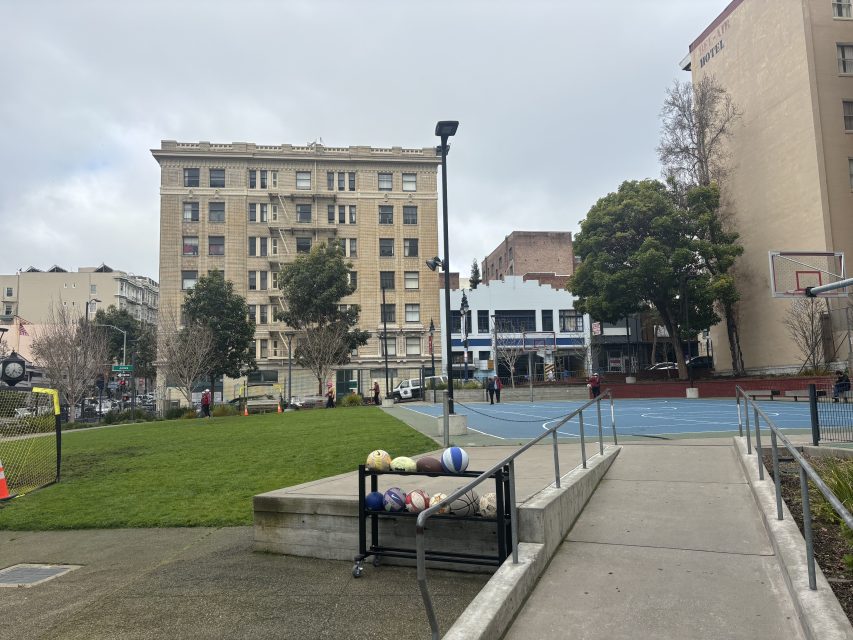A basketball court with a rack of sports balls in the foreground, surrounded by grass, buildings, and a few people on a cloudy day.