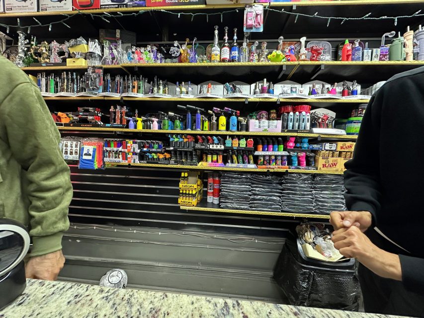 Two people stand at a counter in a shop with shelves behind them displaying lighters, rolling papers, pipes, and smoking accessories.
