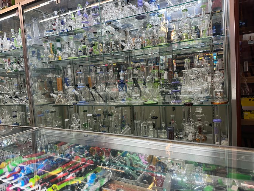 Glass display case filled with various glass water pipes, bongs, and smoking accessories arranged on shelves in a shop, with colorful lighters in the foreground.