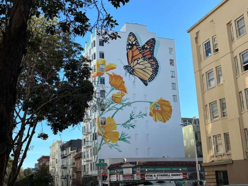A large mural of a monarch butterfly and California poppies covers the side of a white building on a city street corner with nearby trees and other buildings visible.
