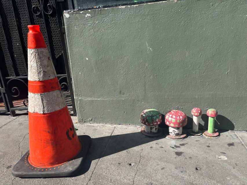 An orange traffic cone stands on a sidewalk next to five small objects painted to look like red and green mushrooms by a grayish-green wall.