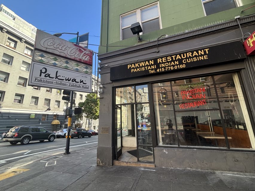 Street view of Pakwan Restaurant, a Pakistani and Indian cuisine eatery with a black and gold sign, glass door, and a sidewalk sign, located at a city intersection.