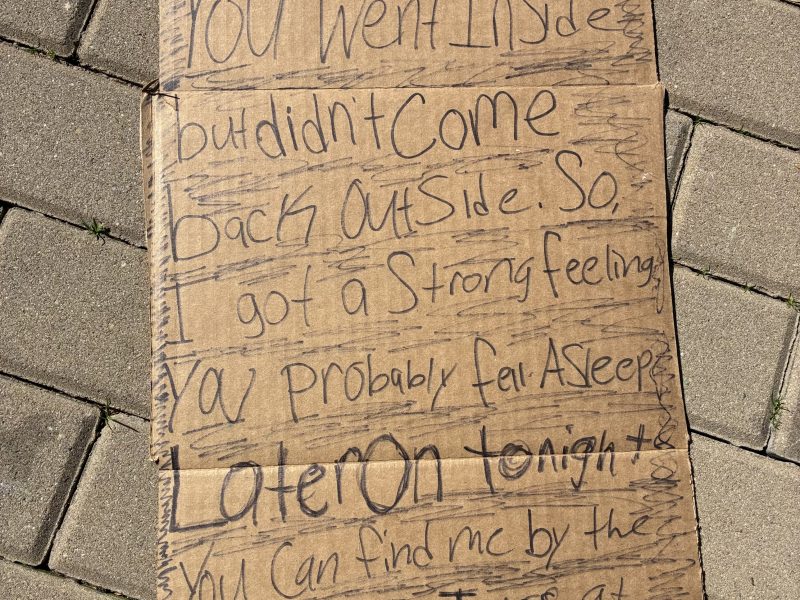 A handwritten message on a cardboard sign lying on pavement, explaining the writer waited outside but didn't see someone return, and can be found later by the same spot near a Samsung S24 ad.