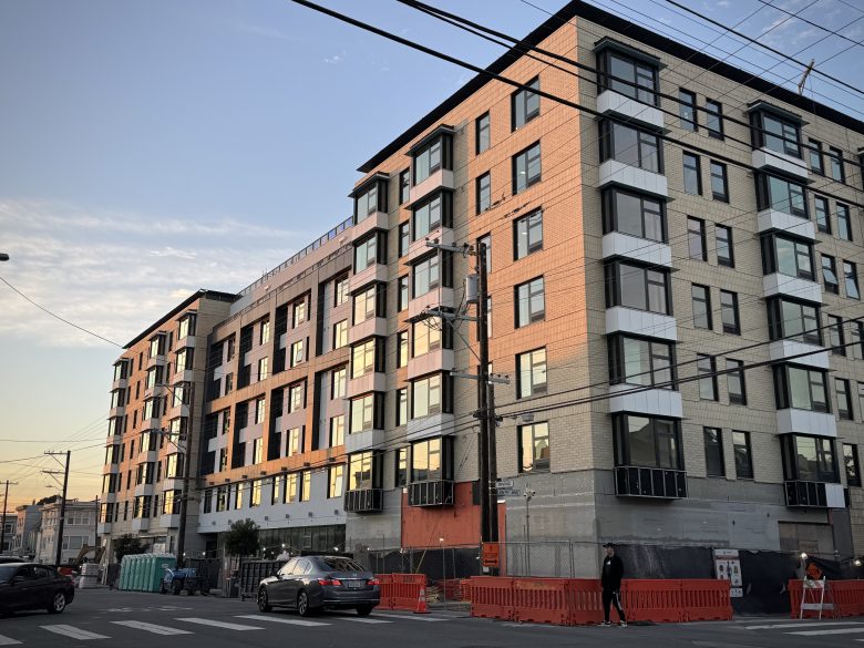 A modern six-story apartment building under construction at a street corner, with orange barriers and construction materials visible at ground level.