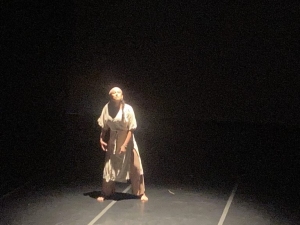 A person in a loose, light-colored outfit stands barefoot and poses under a spotlight on a dark stage.