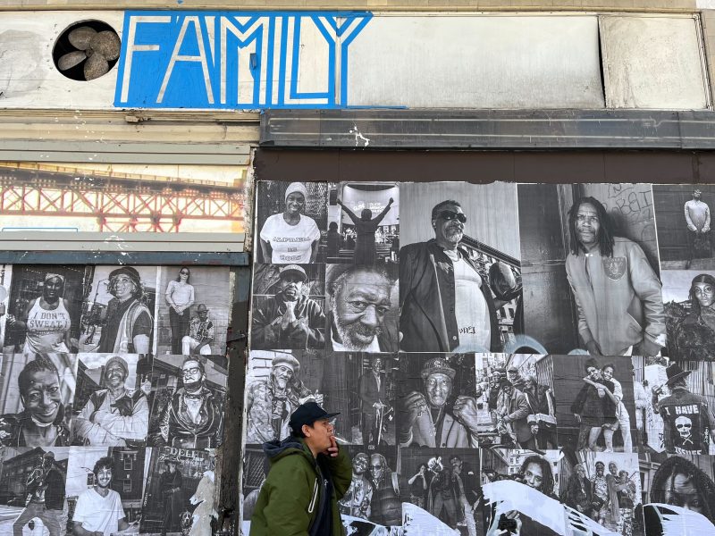 A person walks past a building covered in black-and-white portraits of people, with a blue sign above reading "FAMILY."