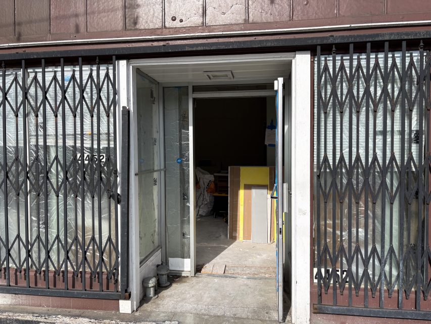 Storefront with metal security gates partially open, glass door ajar, and construction materials visible inside a building under renovation.