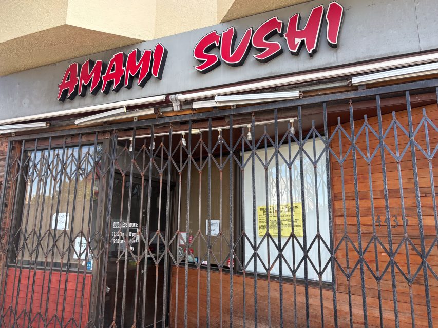 Storefront of Amami Sushi with metal security gate closed and signs posted on the door and window.