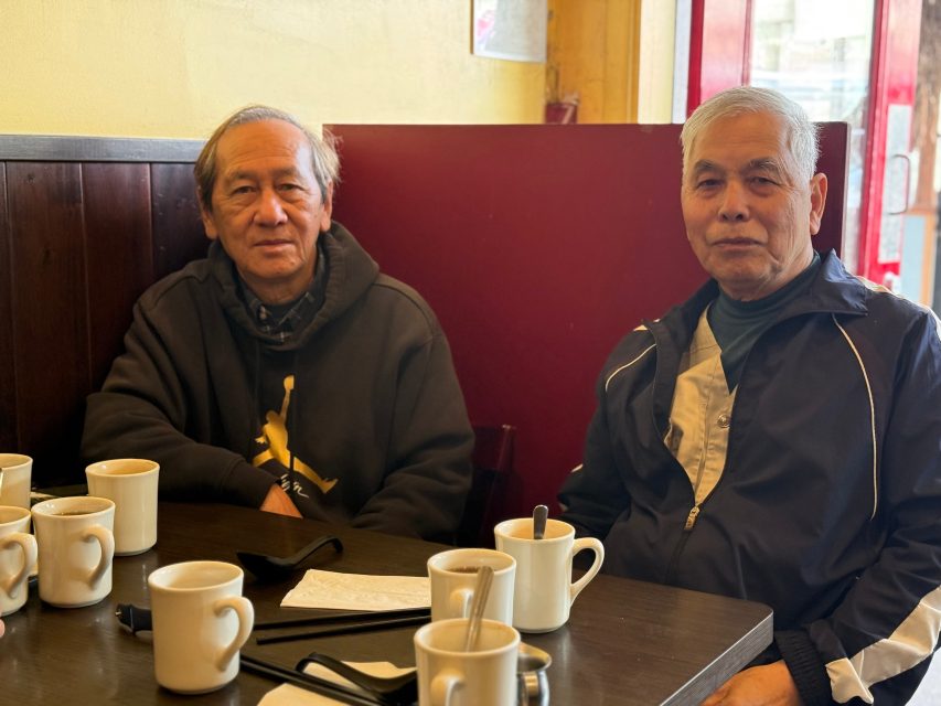 Two older men sit at a table in a cafe with several mugs of coffee, napkins, and utensils in front of them. They are facing the camera and appear relaxed.