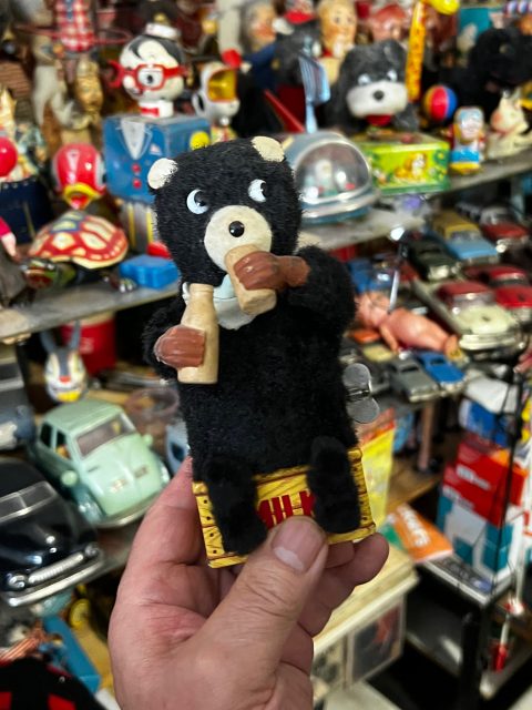 A person holds a vintage black bear toy with a bottle in its paw, surrounded by various retro toys and collectibles on shelves in the background.