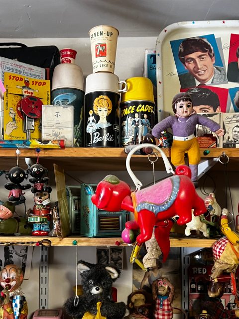A shelf filled with vintage toys, mugs, lunchboxes, figurines, and collectibles, including a red toy horse, a Barbie thermos, and a tray with a portrait of a young man.