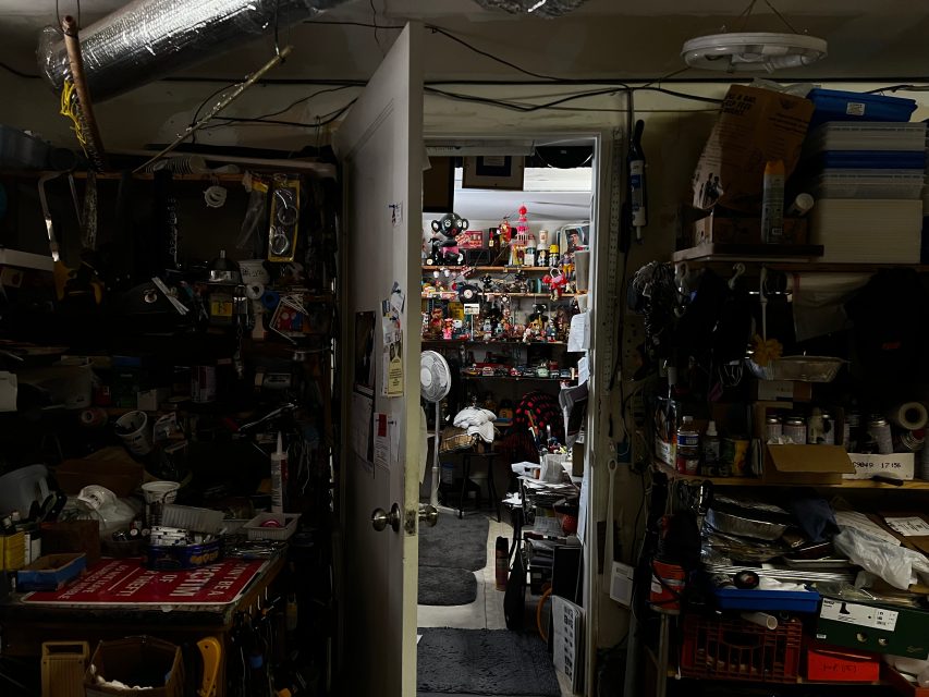A cluttered workshop with shelves full of tools, boxes, and various items. An open door reveals another crowded room filled with collectibles and equipment.