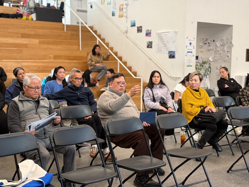 A group of adults sits in folding chairs facing forward; one man in the center gestures as he speaks. Others listen attentively in a bright room with stairs and posters on the wall.