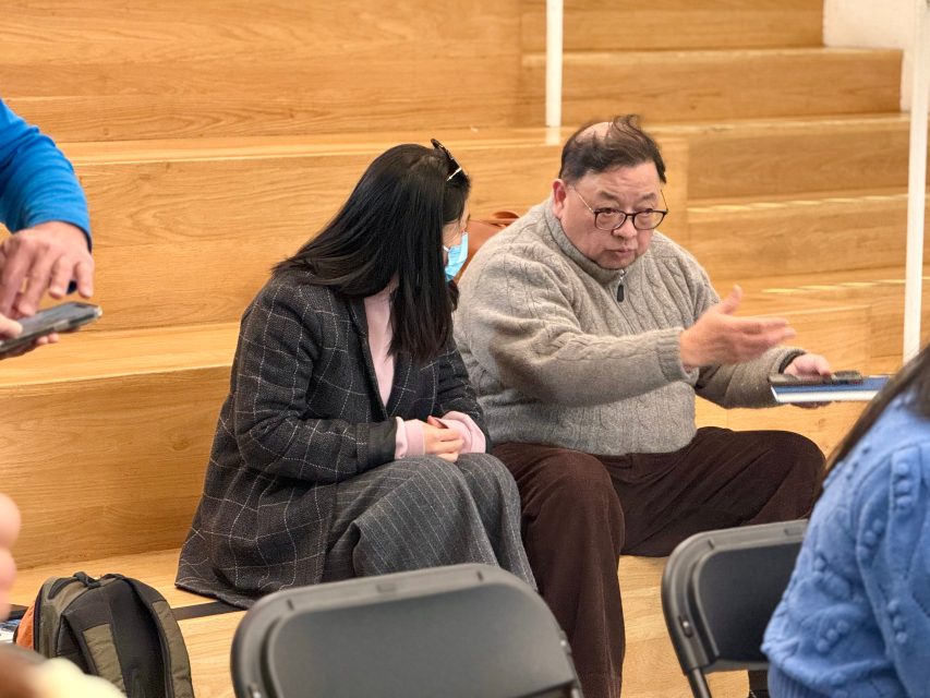Two people sit on wooden bleachers having a conversation; one gestures with his hand while the other listens and holds a notebook.