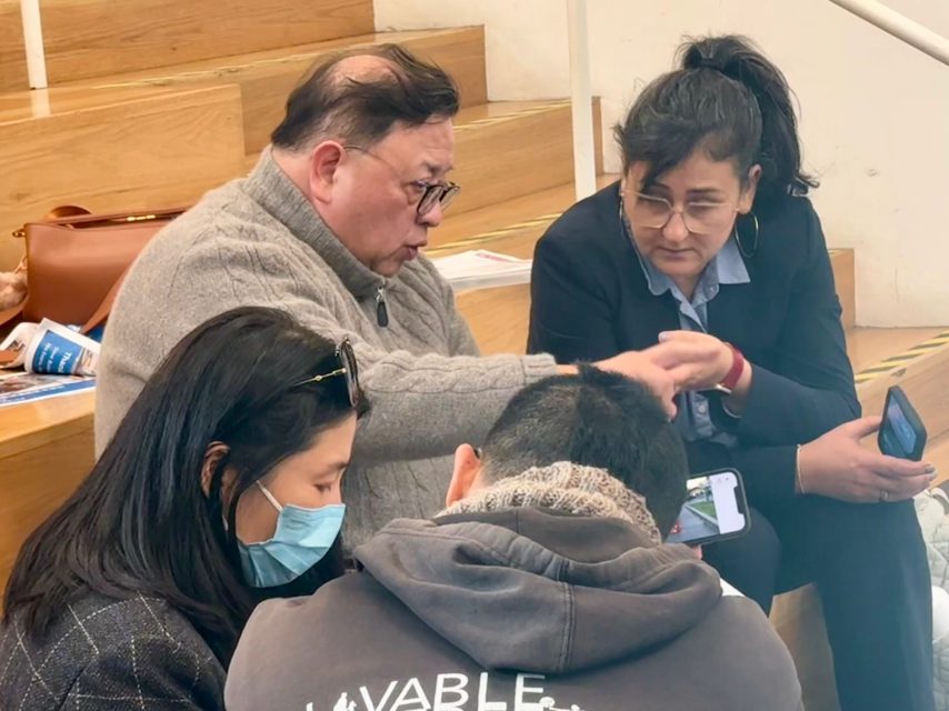 Four people sit together, two facing forward and two in profile, engaged in conversation; one person wears a mask, and two hold smartphones.