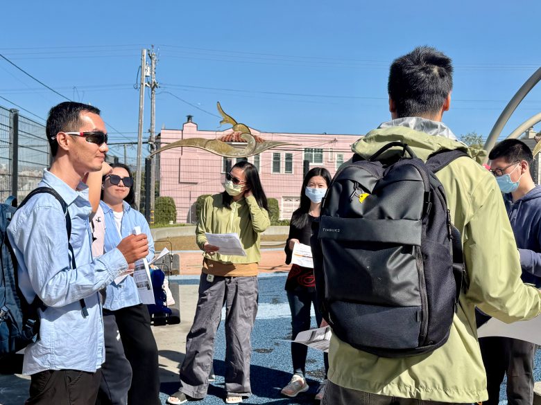 A group of people stand outdoors in sunlight, some holding papers and wearing masks, engaged in conversation near a pink building.