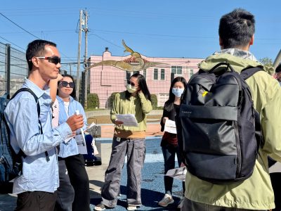 A group of people stand outdoors in sunlight, some holding papers and wearing masks, engaged in conversation near a pink building.
