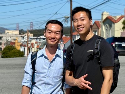 Two men with backpacks stand on a sunny street with houses and power lines in the background; the Golden Gate Bridge is visible in the distance.