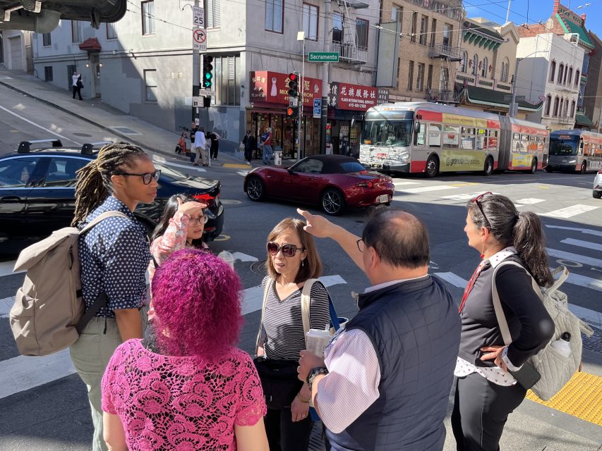 A group of six people stand at a crosswalk in a city, engaged in conversation near a street corner with shops, cars, and a bus in the background.
