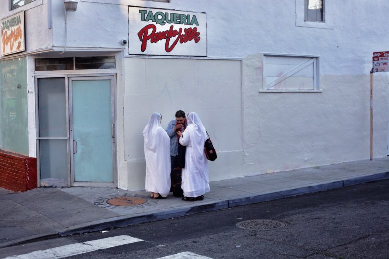 Two people in white robes stand with a man on a city sidewalk in front of a building with a "Taqueria" sign.