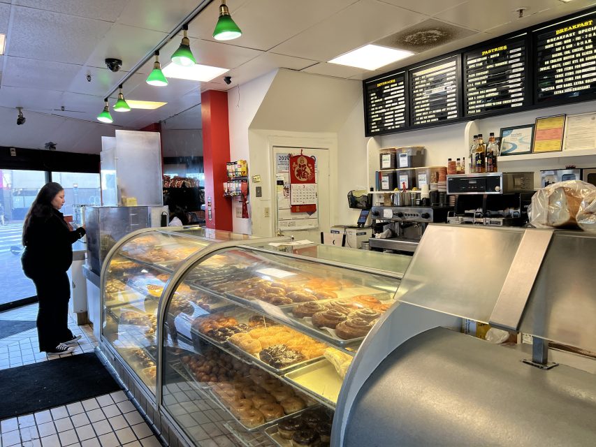 A woman stands at the counter of a bakery or donut shop, with a glass display case filled with assorted pastries and a menu board overhead.