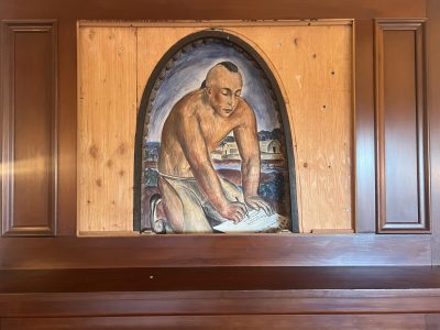 A painting of a shirtless man working is partially framed by unfinished wood panels within a wooden wall.