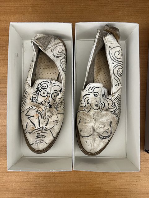 A pair of white slip-on shoes in boxes, each decorated with black line art portraits of a man on the left shoe and a woman on the right shoe.