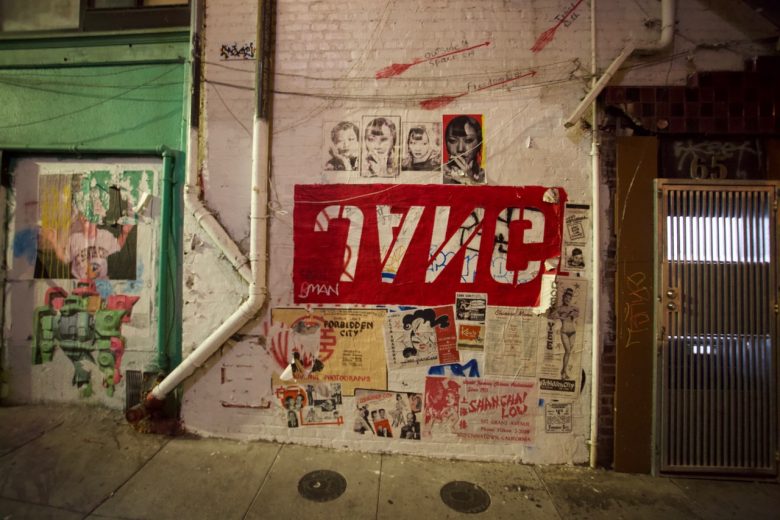 Wall with various posters, graffiti, and flyers, including a large red sign with mirrored text, on a city street corner near a metal door.
