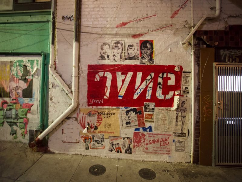 A brick wall with posters, graffiti art, and a large red and white sign reading โSYNCโ in reverse, located in an urban alley.