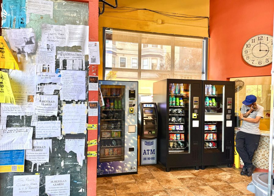 Person standing by three vending machines and an ATM in a brightly lit room with an orange wall and clock; a bulletin board covered with handwritten notes is visible on the left.