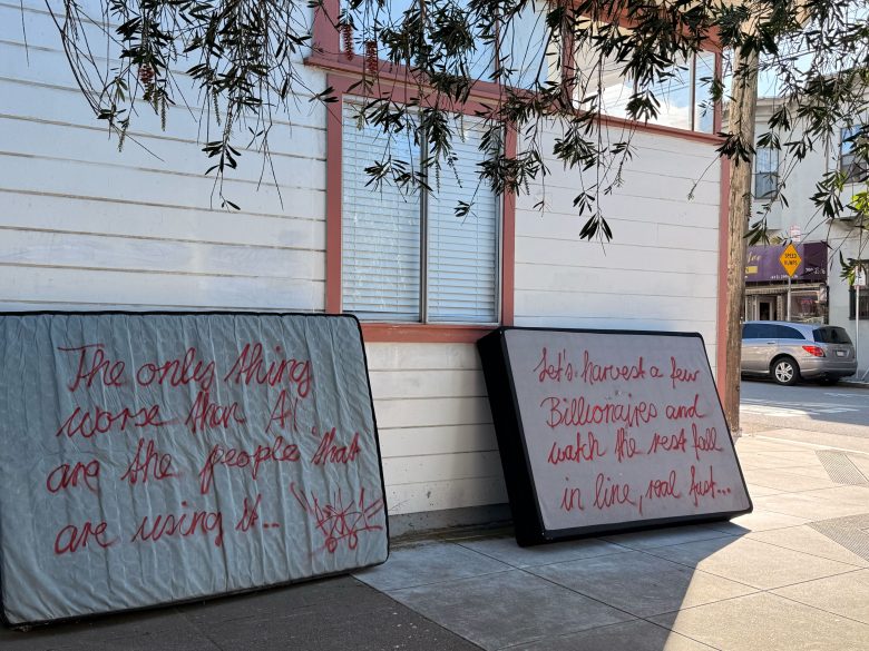 Two mattresses with red handwritten text lean against a building on a city sidewalk; one mattress partially blocks a window. Trees and street signs are visible nearby.
