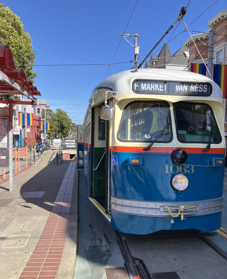 A vintage blue and cream streetcar marked "F Market & Van Ness" is stopped at a city tram station with rainbow flags displayed on buildings nearby.
