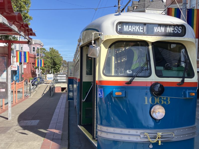 A vintage blue and cream streetcar marked "F Market & Van Ness" is stopped at a city tram station with rainbow flags displayed on buildings nearby.